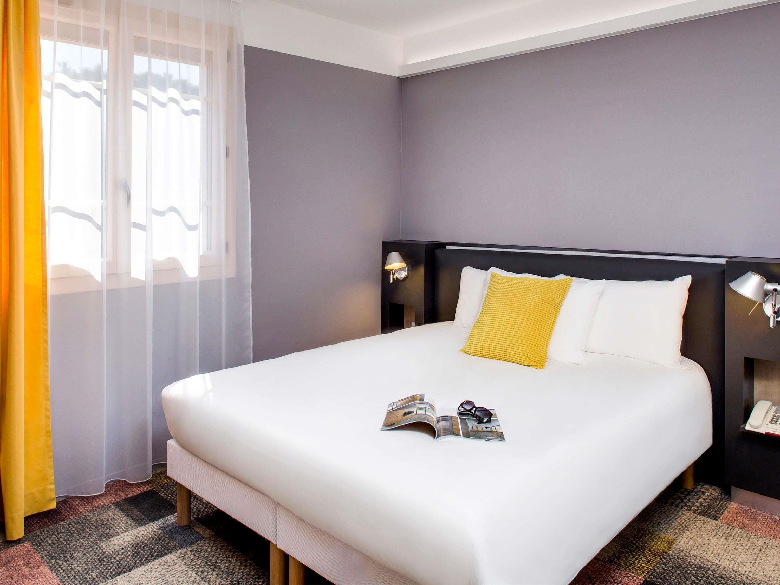A hotel room with a large bed, a bedside table with a lamp, a window with sheer curtains, and a view of the outside.