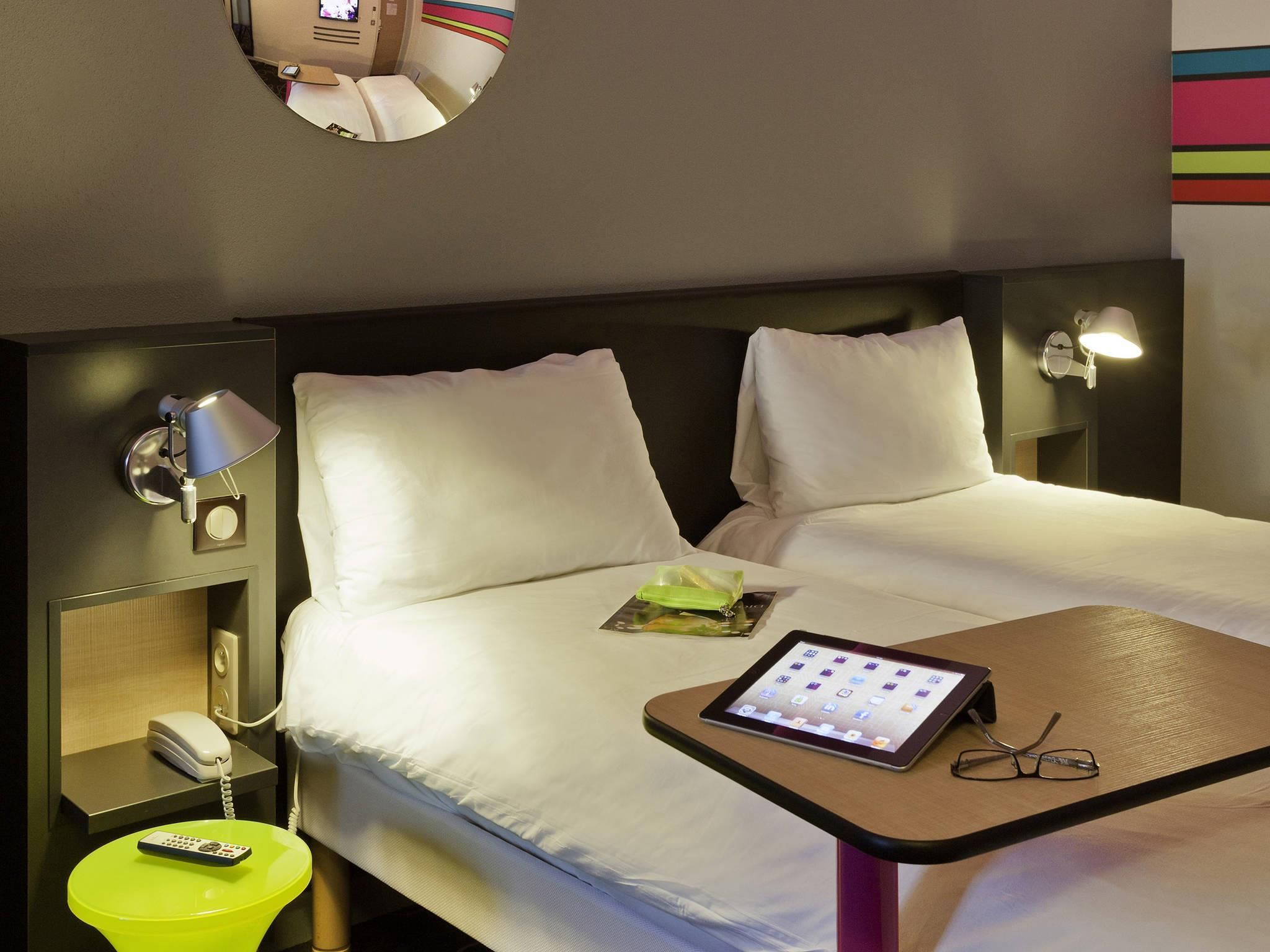 Ein Hotelzimmer mit zwei Betten, einem Nachttisch mit einem Tablet und Gläsern, einem Telefon auf einem Ständer und einem Spiegel über dem Bett.