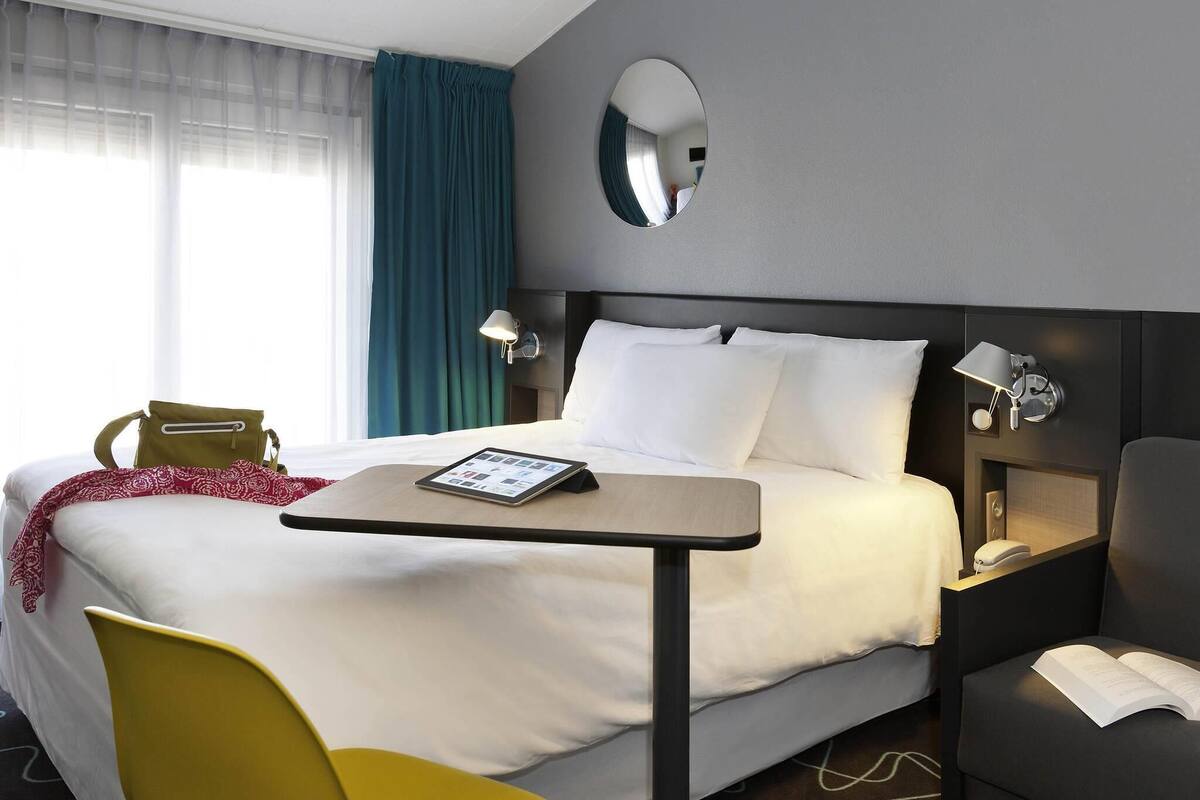 A hotel room with a bed, bedside tables, a chair, a desk, a mirror, and a window with curtains.