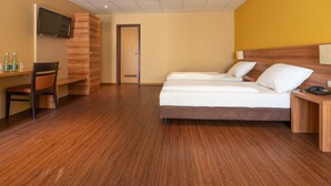 A hotel room with a wooden floor, a bed with white linens, a desk with a chair, a television mounted on the wall, and a door leading to another room.