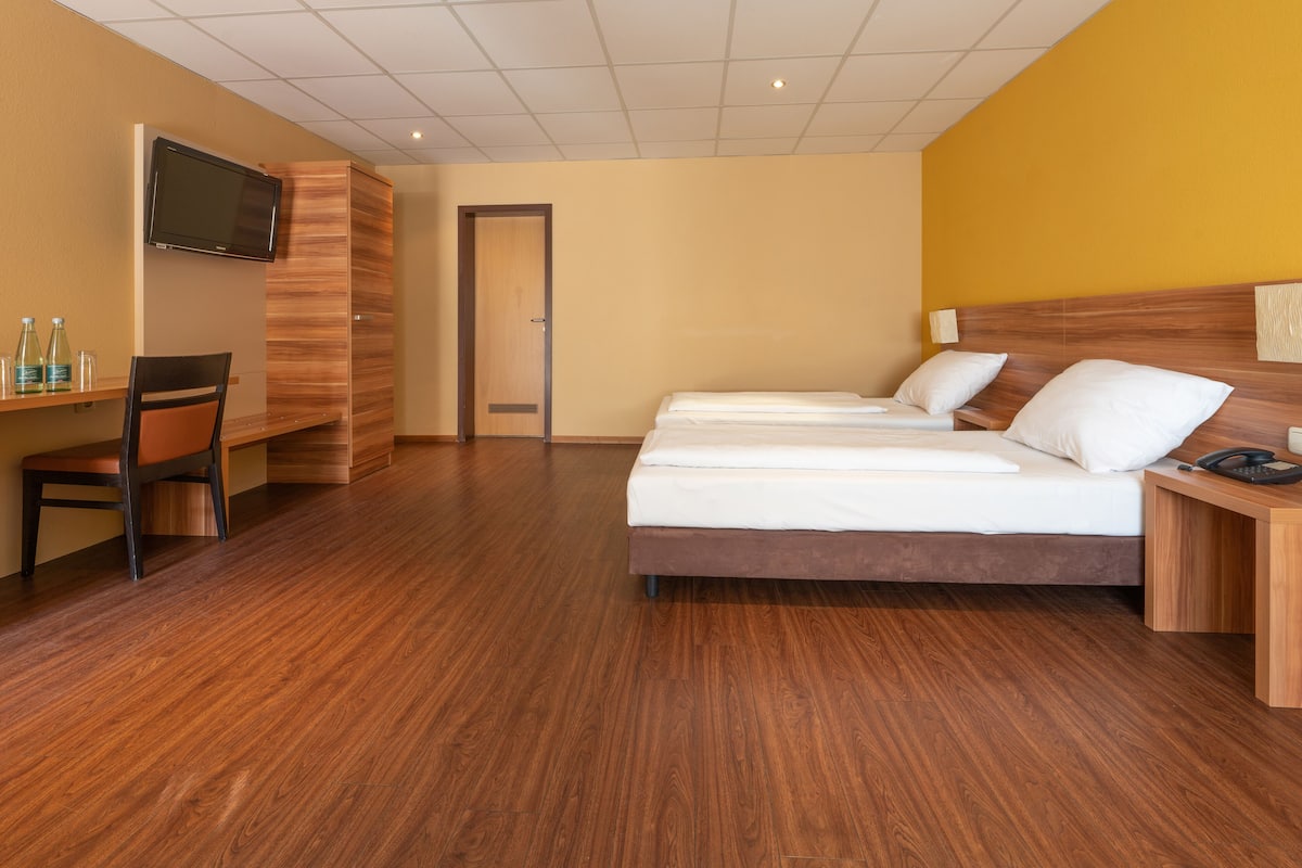 A hotel room with a wooden floor, a bed with white linens, a desk with a chair, a television mounted on the wall, and a door leading to another room.