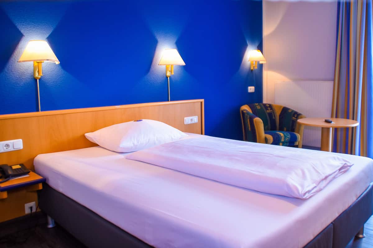 A hotel room with a bed, two wall-mounted lamps, a chair, a small table, and a telephone on the nightstand.