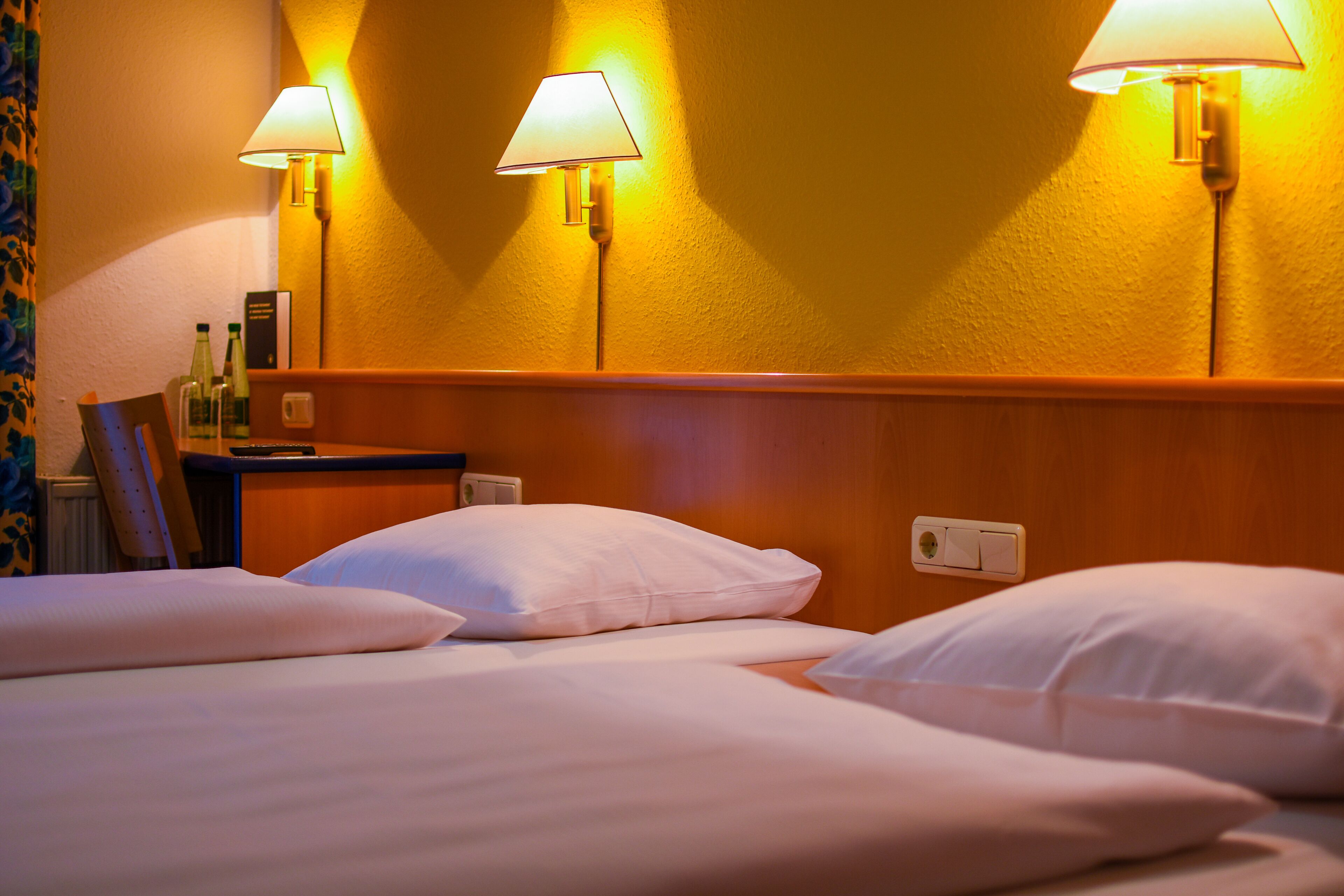 A hotel room with two beds, a nightstand, and two wall-mounted lamps.