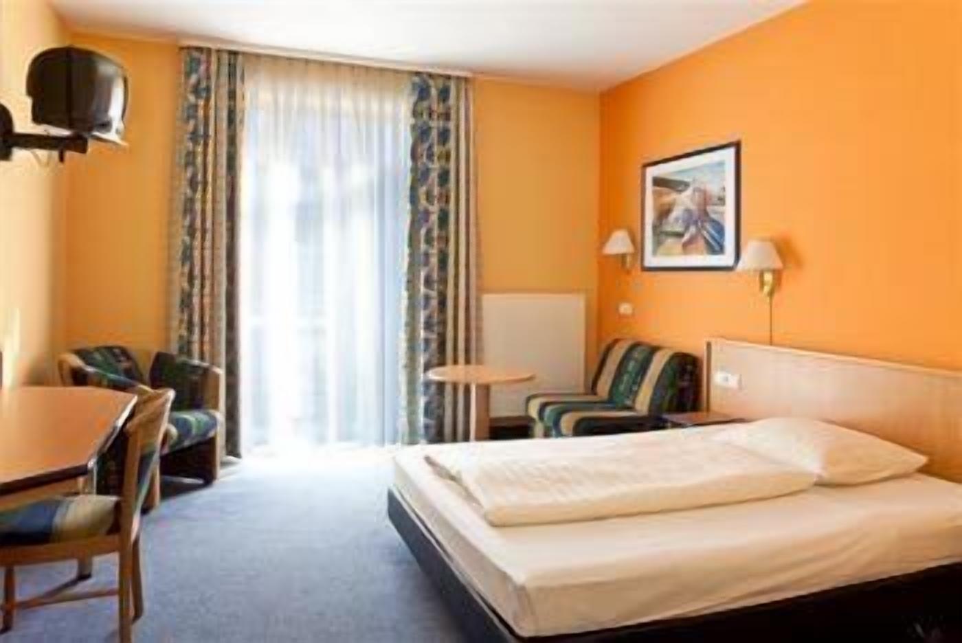 Una habitación de hotel con una cama, un escritorio, dos sillas, una mesita y un ventanal con cortinas.