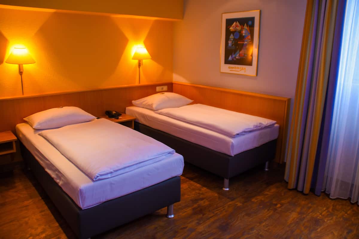 A hotel room with two single beds, wooden flooring, and two wall-mounted lamps.