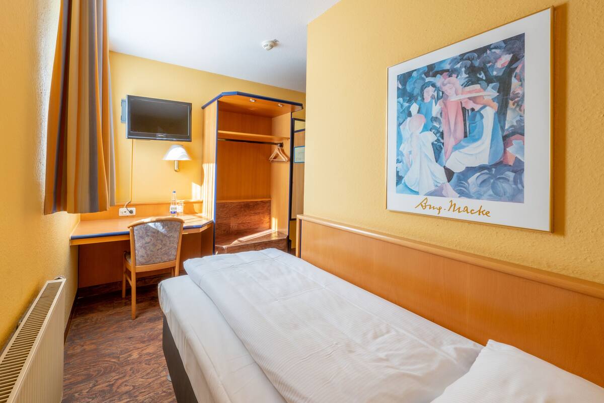 A hotel room with a bed, a desk with a chair, a television mounted on the wall, and a framed picture.