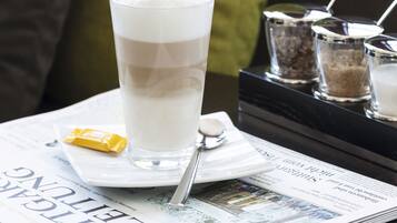 Un verre contenant une boisson claire est posé sur un plateau, accompagné d’une cuillère, d’un objet jaune et d’un journal. La scène se déroule sur une table, avec un autre plateau noir et des coussins verts en arrière-plan.