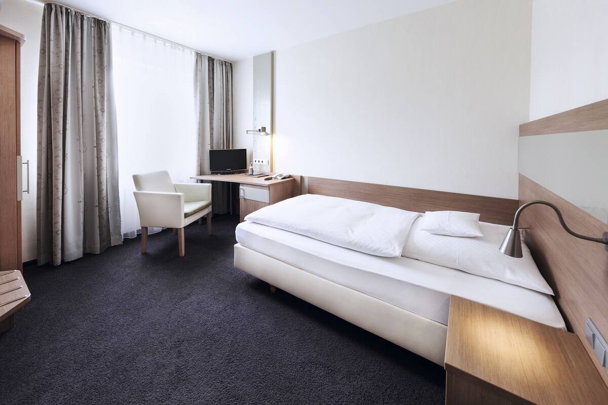 A hotel room with a bed, a desk with a computer, a chair, and a large mirror.