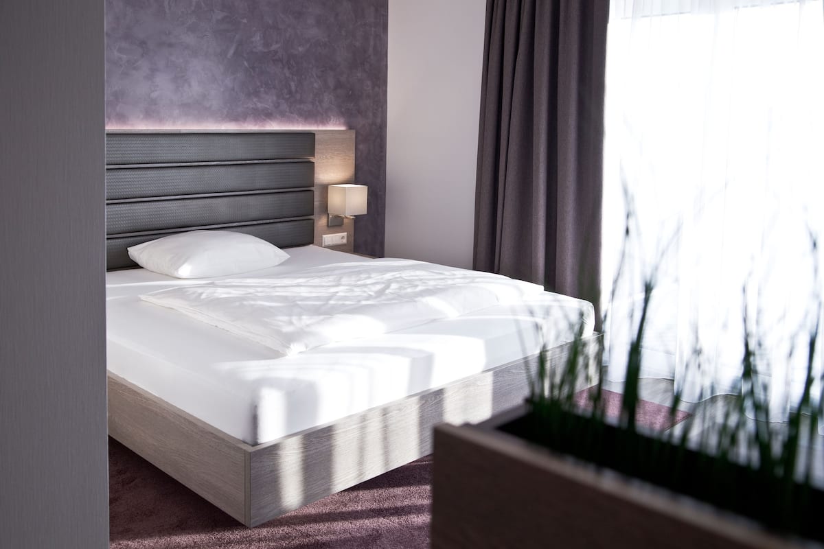 A modern hotel room with a large bed, a headboard with built-in lighting, a bedside table with a lamp, and a view of trees through the window.