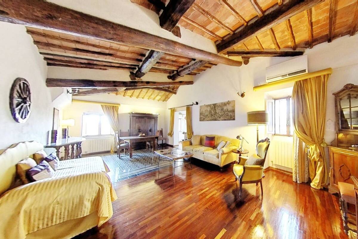 A spacious room with wooden beams on the ceiling, a large window with curtains, a sofa, a table with a lamp, and a wooden cabinet.