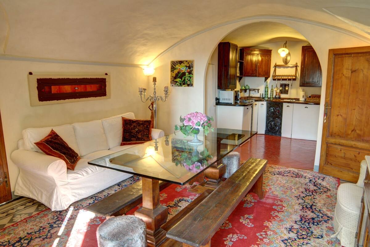 A living room with a glass coffee table, white sofa, and a bench. There is a kitchen in the background with wooden cabinets and a microwave.