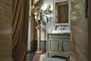 A bathroom with a marble floor, a green vanity with a mirror, and a vase of flowers on a pedestal. - Palazzo Coli Bizzarrini (Siena)