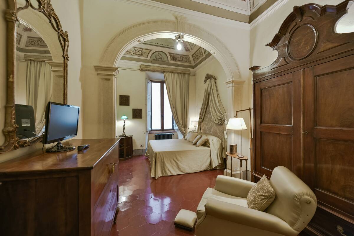 A hotel room with a bed, a desk with a computer, a chair, and a large wooden wardrobe.