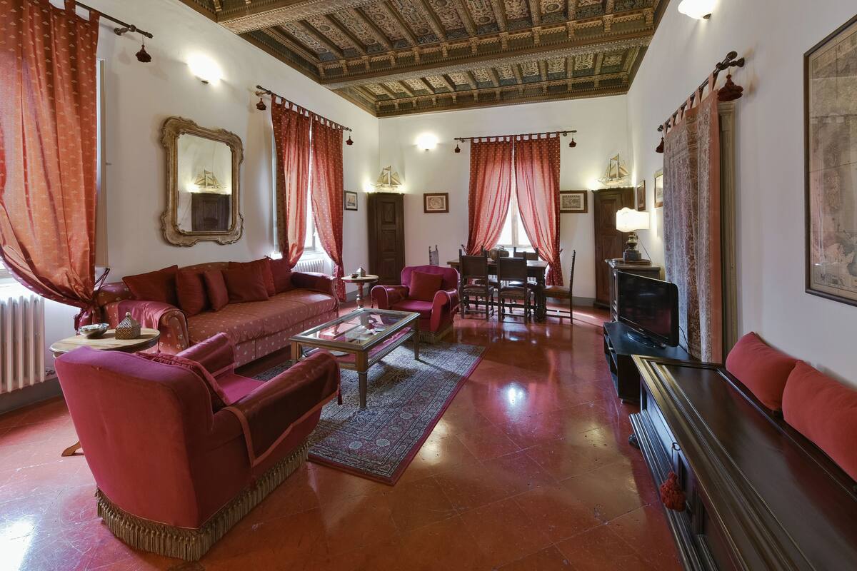 A spacious living room with a red sofa, a coffee table, and a television. There is a dining area with a table and chairs, and a large mirror on the wall.