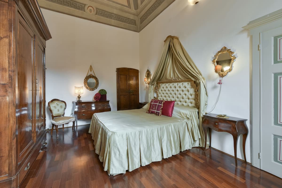 A bedroom with a large bed, a wooden wardrobe, a chair, a nightstand, and a mirror.