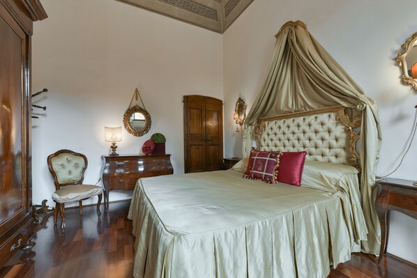 A bedroom with a large bed, a canopy, a wooden wardrobe, a chair, a dresser, and a mirror.