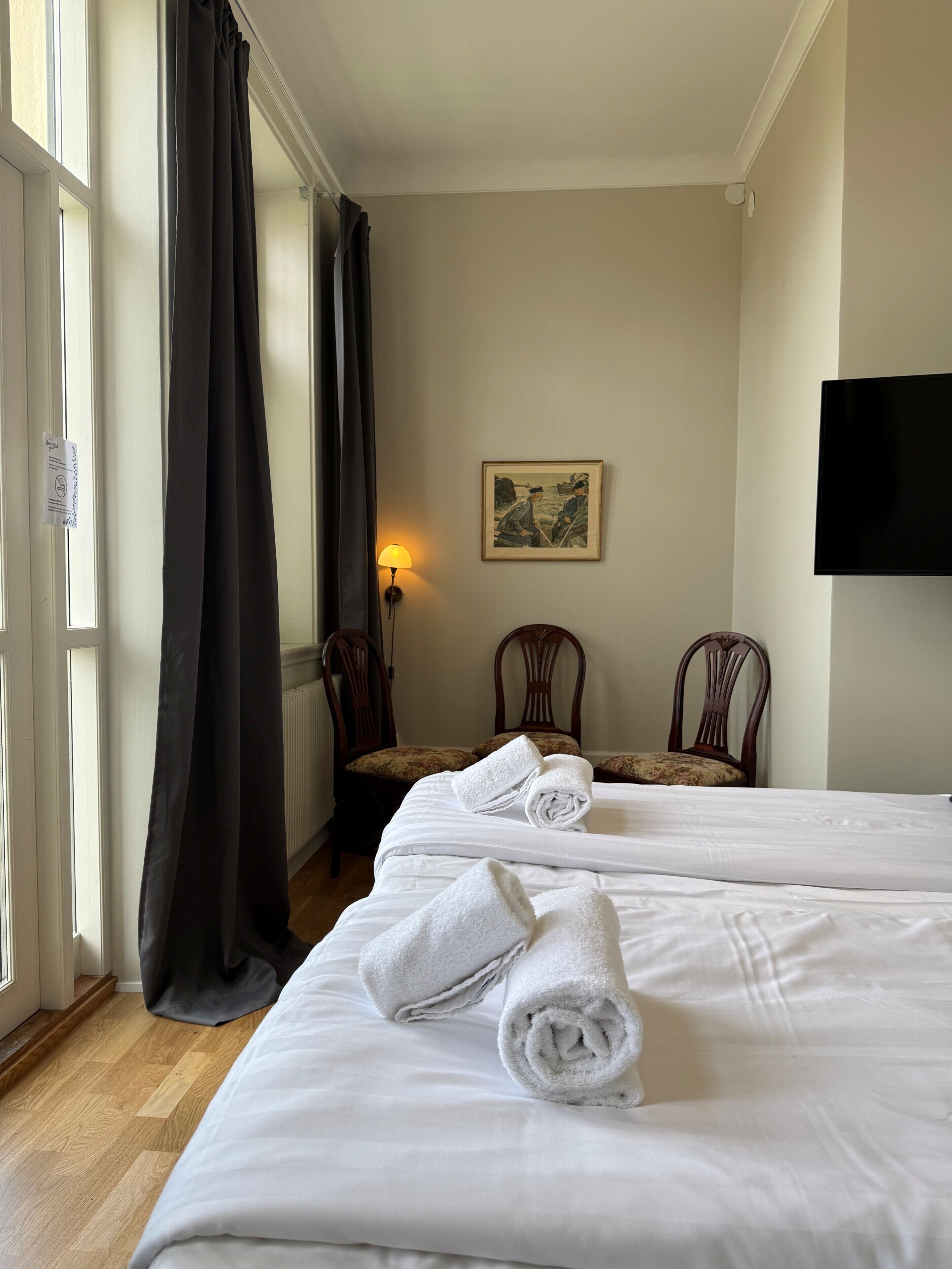 A hotel room with two beds, a painting on the wall, a TV mounted above the beds, and a window with curtains.
