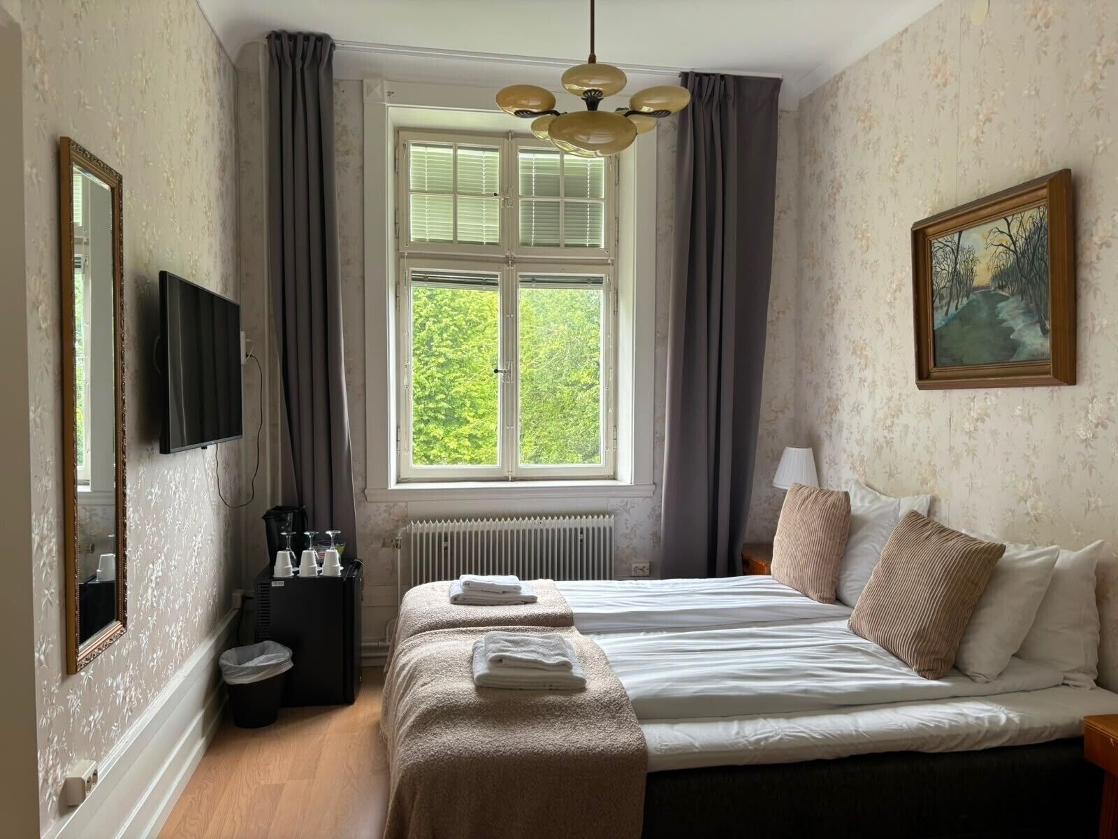 A hotel room with a bed, a television, a window with curtains, a radiator, and a painting on the wall.