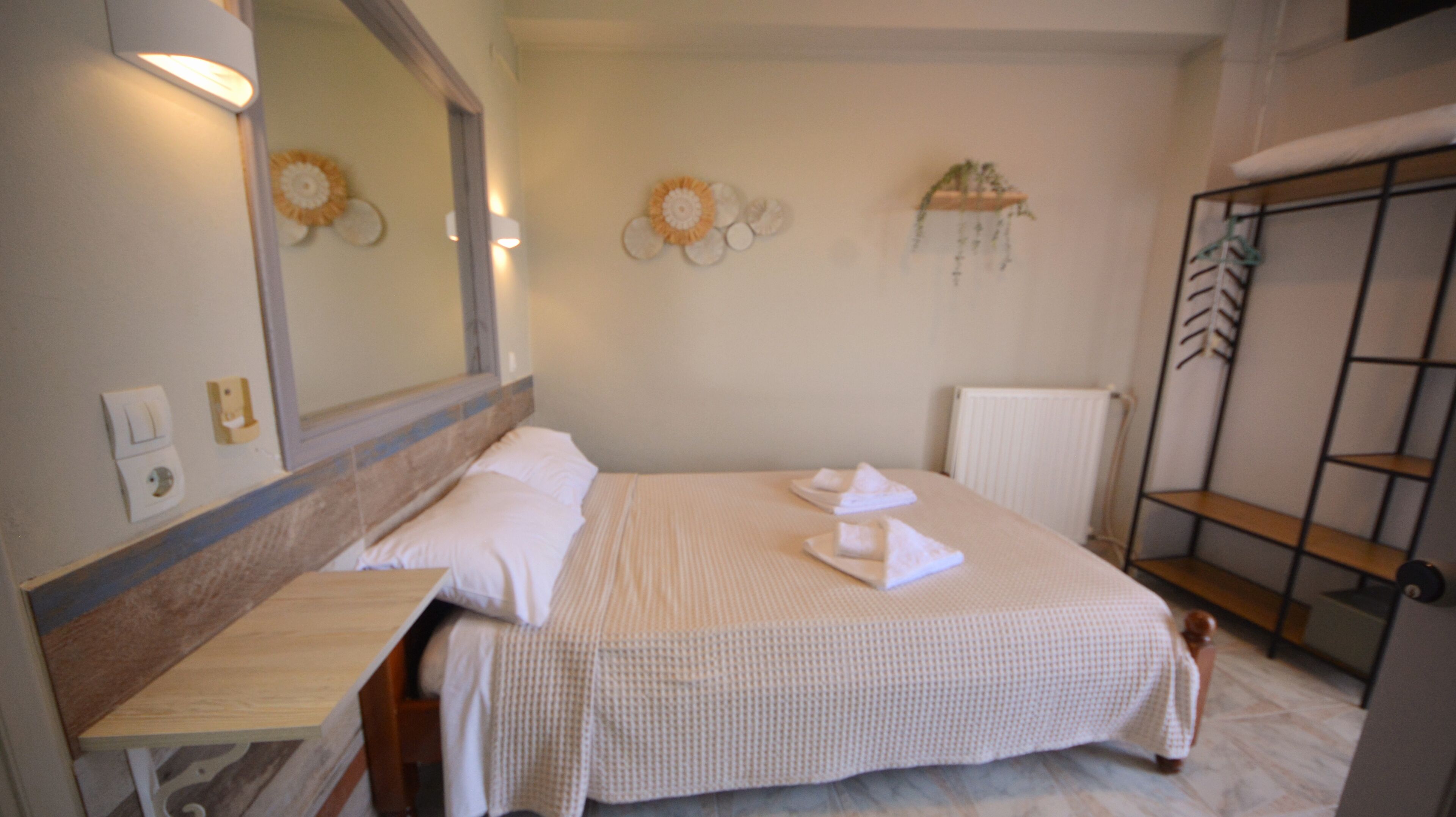 A hotel room with a bed, bedside table, mirror, wall decor, radiator, and a shelf unit.