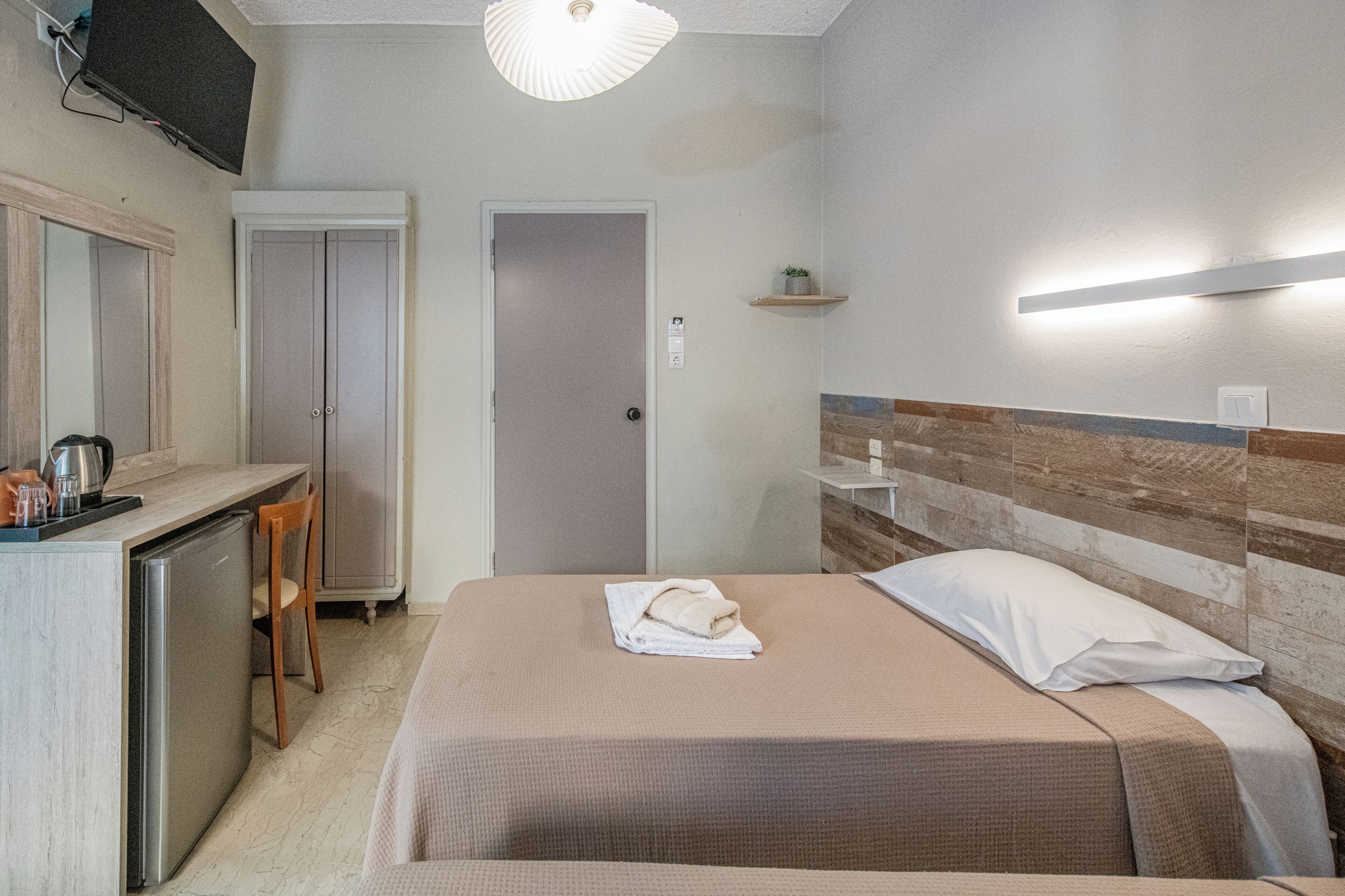 A hotel room with a bed, a desk, a chair, a wardrobe, and a small shelf with a plant.