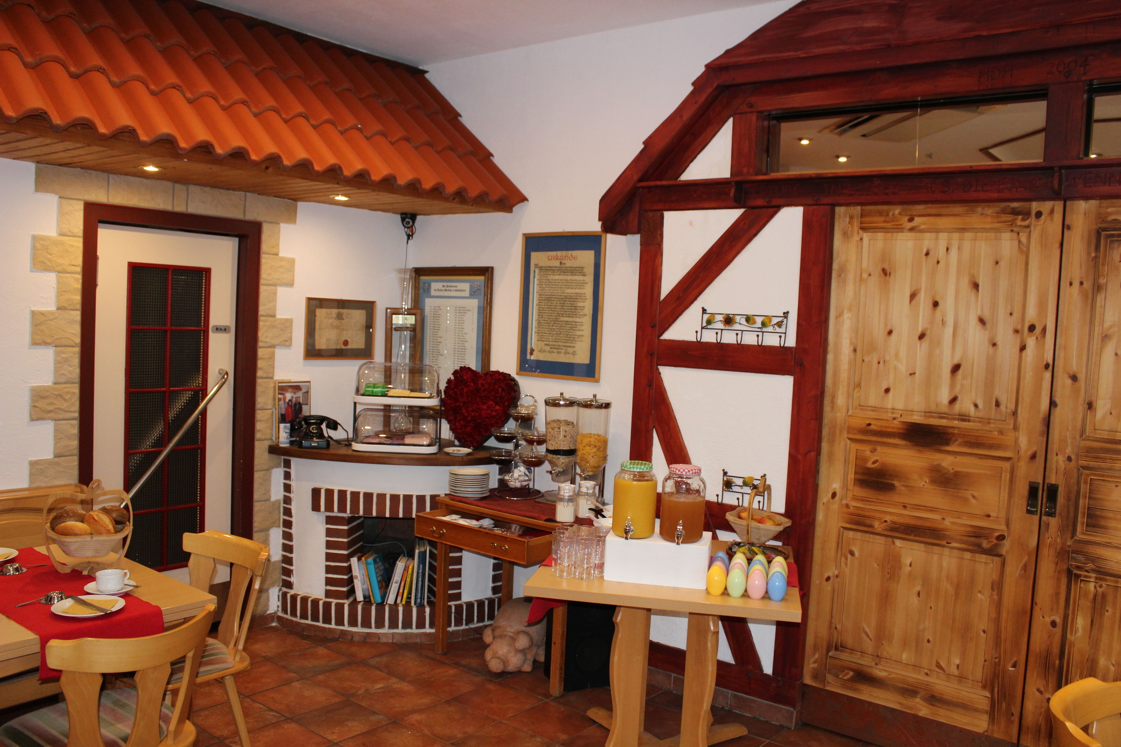 A cozy dining area with a wooden table, chairs, and a red tablecloth. There is a fireplace with a brick surround and a wooden bench.
