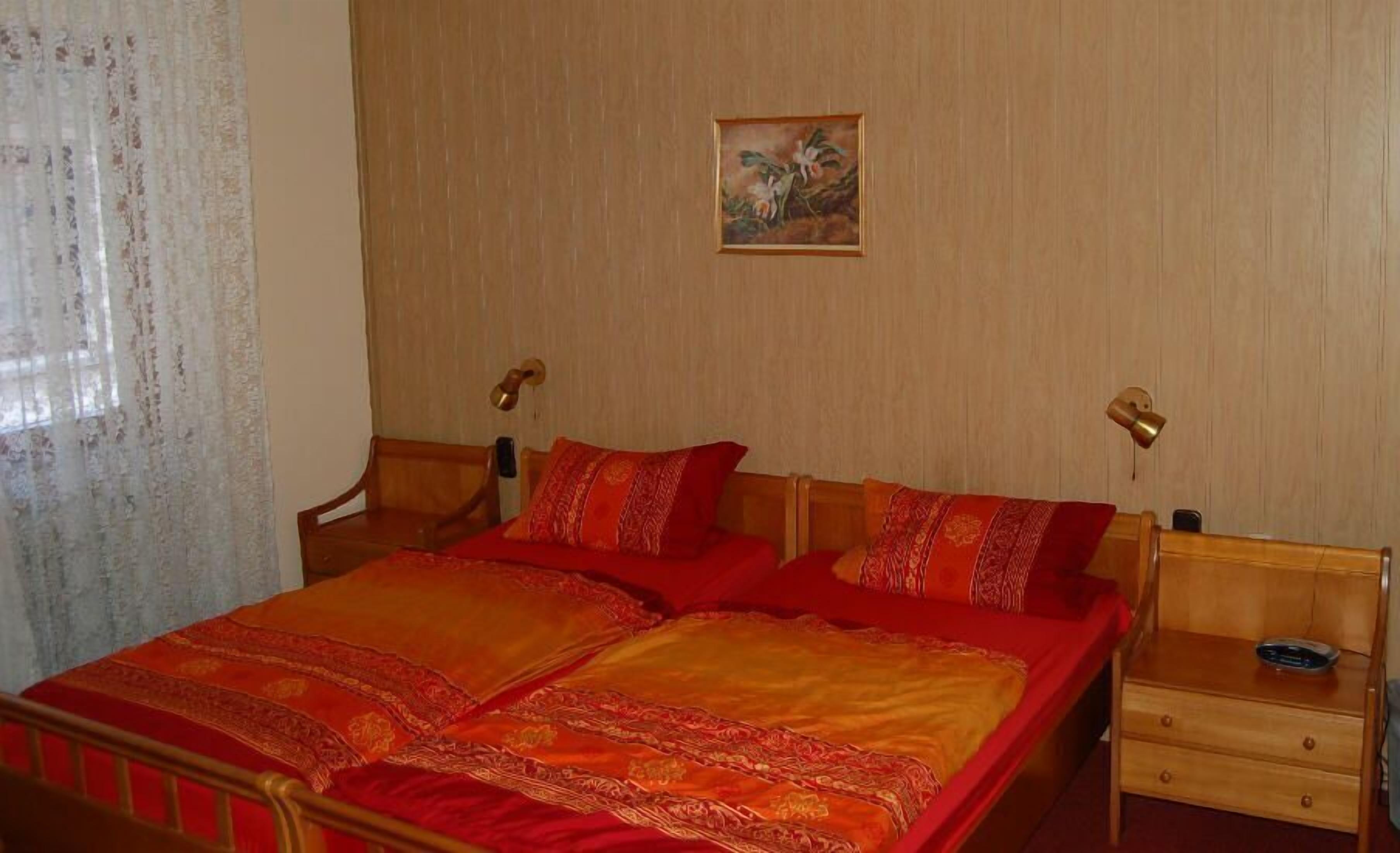 A room with a bed, two bedside tables, a small painting on the wall, and a window with curtains.