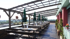 An outdoor patio with wooden tables and benches, green umbrellas, and potted plants. - Hotel Panon (Hodonin)