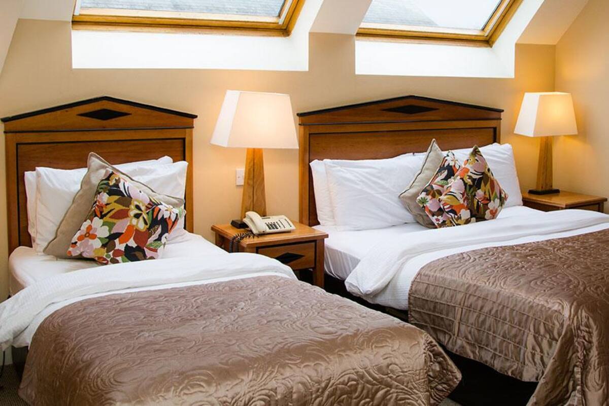 Une chambre d’hôtel avec deux lits, chacun doté d’une tête de lit en bois, de draps blancs et d’un coussin à motifs floraux colorés. On y trouve des lampes de chevet et un téléphone sur la table de chevet. La chambre est éclairée par des puits de lumière.