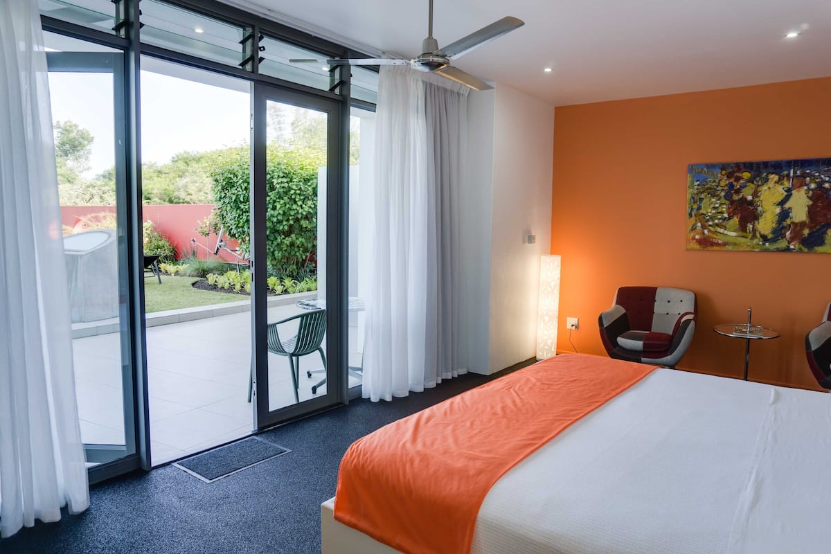 Hotelkamer met een bed, twee stoelen, een kleine tafel, een plafondventilator en een glazen schuifdeur naar een buitenruimte met terras en groen.