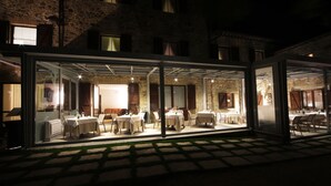 Een terras met tafels en stoelen, een stenen gebouw op de achtergrond, en een glazen constructie die het terras van de straat scheidt.
