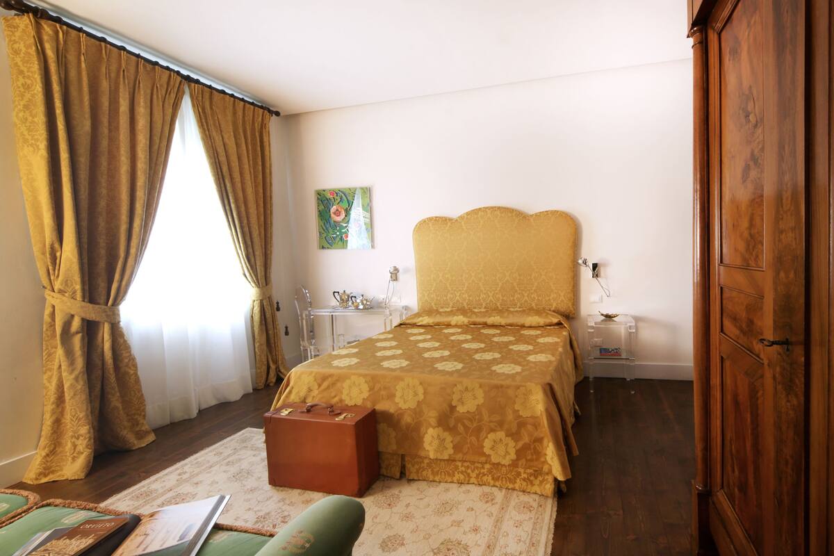 A bedroom with a bed, a wooden wardrobe, a small table with a lamp, and a suitcase on the floor.