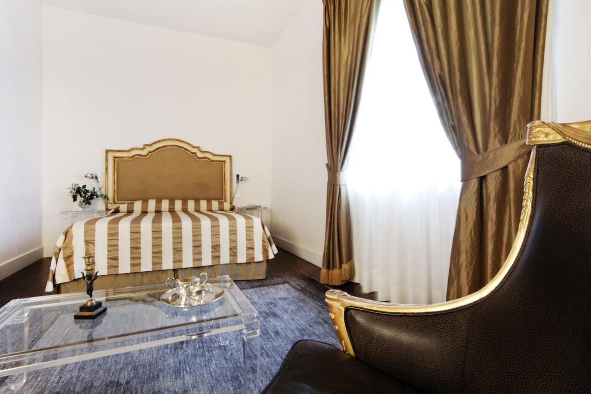 A hotel room with a bed, a clear glass coffee table, a dark leather armchair, and a window with curtains.