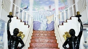 Un grand escalier orné d’une fresque murale, flanqué de deux statues noires de chaque côté, et doté de rampes blanches.