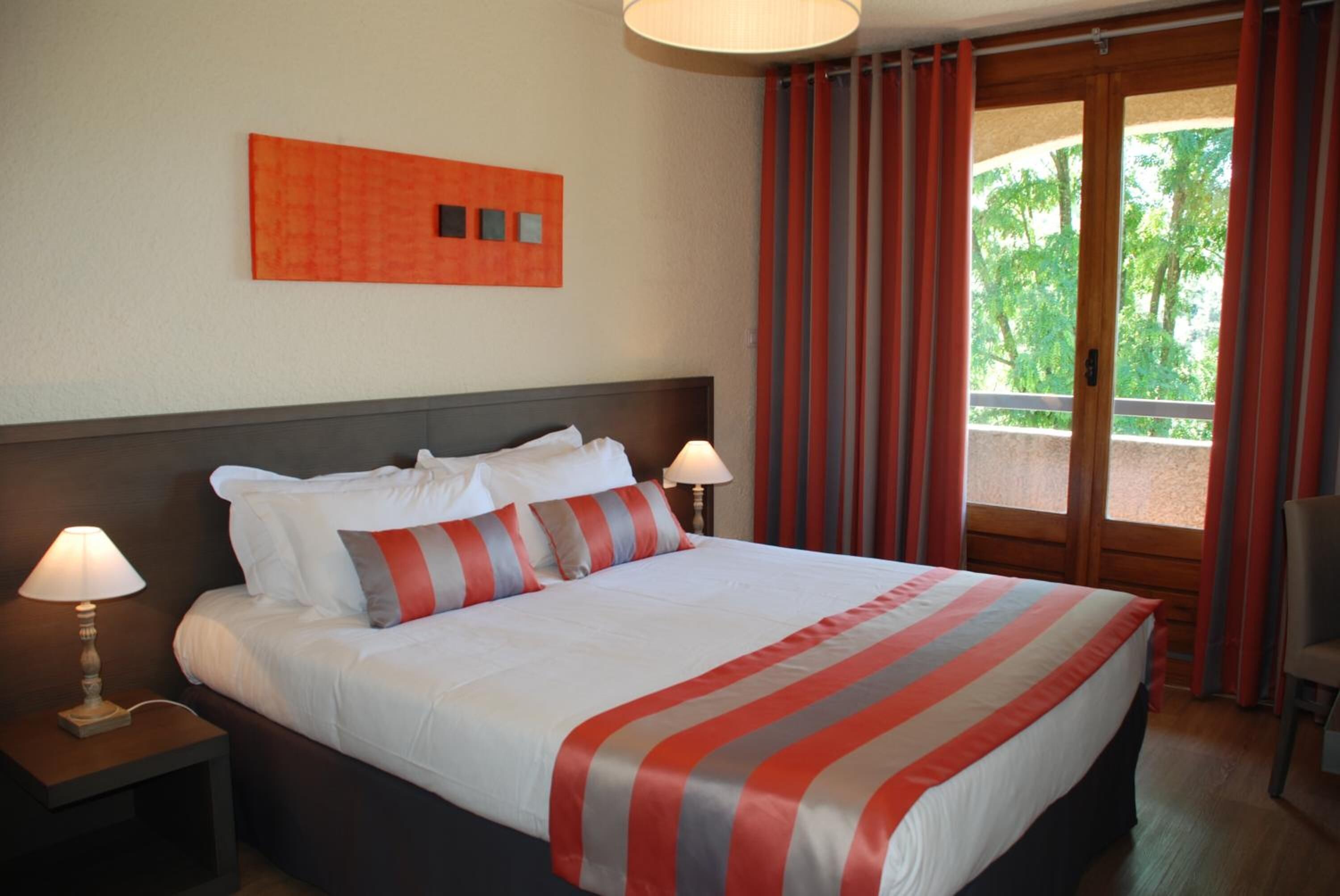 A hotel room with a large bed, two bedside lamps, a view of greenery through the window, and a red-striped throw blanket.