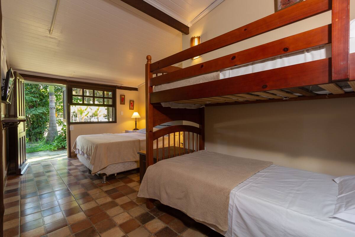 A room with two bunk beds, a checkered floor, a window with a view of greenery, and a lamp.