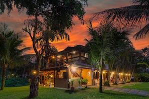 Um edifício de dois andares com varanda, rodeado por palmeiras e um gramado bem cuidado.