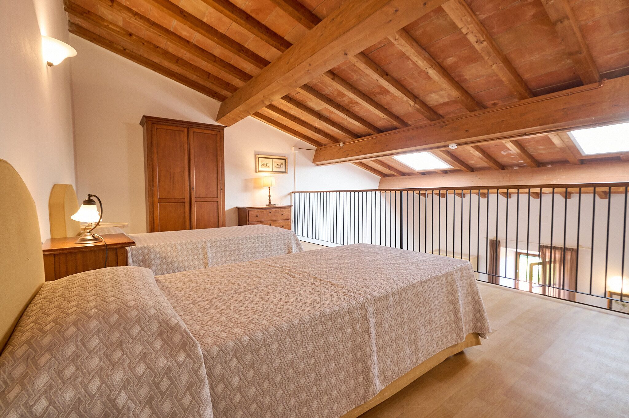 A bedroom with two beds, wooden beams, a balcony with a railing, and a small table with a lamp.