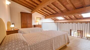 A bedroom with two beds, wooden beams, a balcony with a railing, and a small table with a lamp.