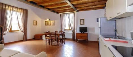 A spacious living area with a wooden ceiling, a dining table with chairs, a sofa, a television, and a kitchen with white cabinets and a sink.