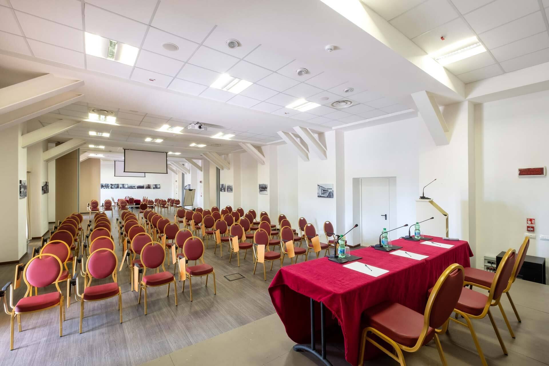 A conference room with rows of red chairs, a long table with a red tablecloth, and a projector screen at the front.