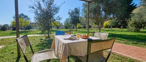 An outdoor dining area with a table and chairs under a pergola, surrounded by grass and trees.