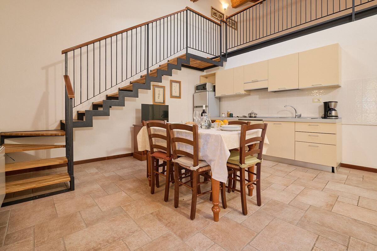 Un coin repas avec une table dressée pour quatre personnes, un escalier et une cuisine en arrière-plan.