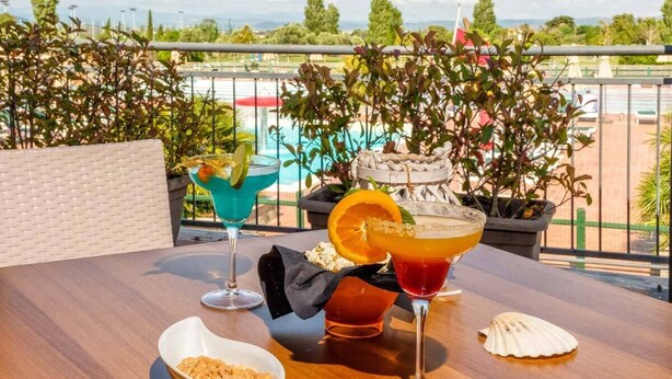 Een tafel met twee drankjes, een schaal met snacks en een decoratieve schelp op een balkon met uitzicht op een zwembad.