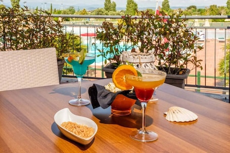 A table with two drinks, a bowl of snacks, and a decorative shell on a balcony overlooking a pool.