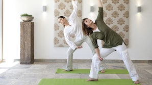 Duas pessoas praticando ioga em tapetes verdes num quarto com papel de parede estampado e um pilar de madeira com uma planta.
