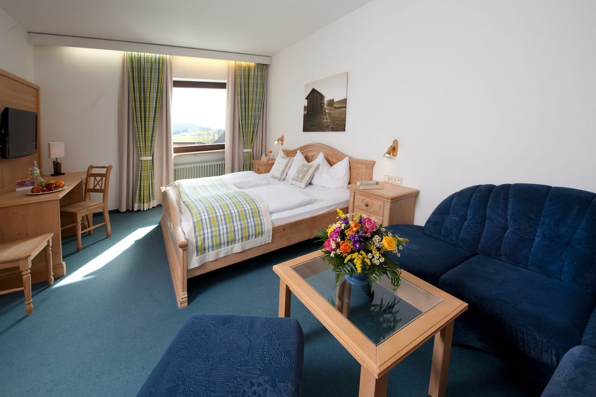 Une chambre d’hôtel avec un lit, un canapé bleu, un bureau en bois, une chaise, une petite table avec un vase de fleurs et une fenêtre avec des rideaux.