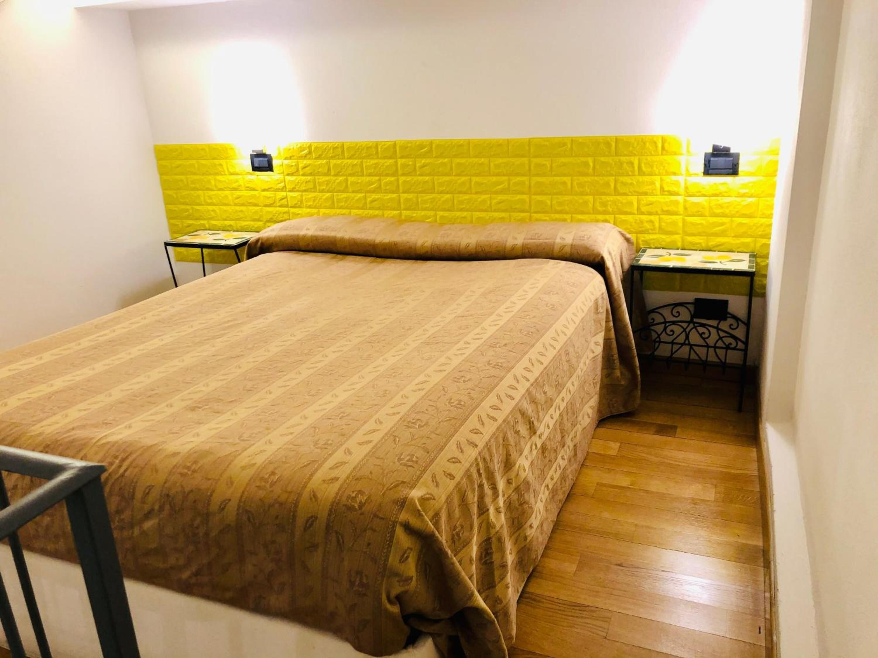 Una cama individual con una colcha estampada en una habitación con pared de ladrillo amarillo y una pequeña mesa con una silla.