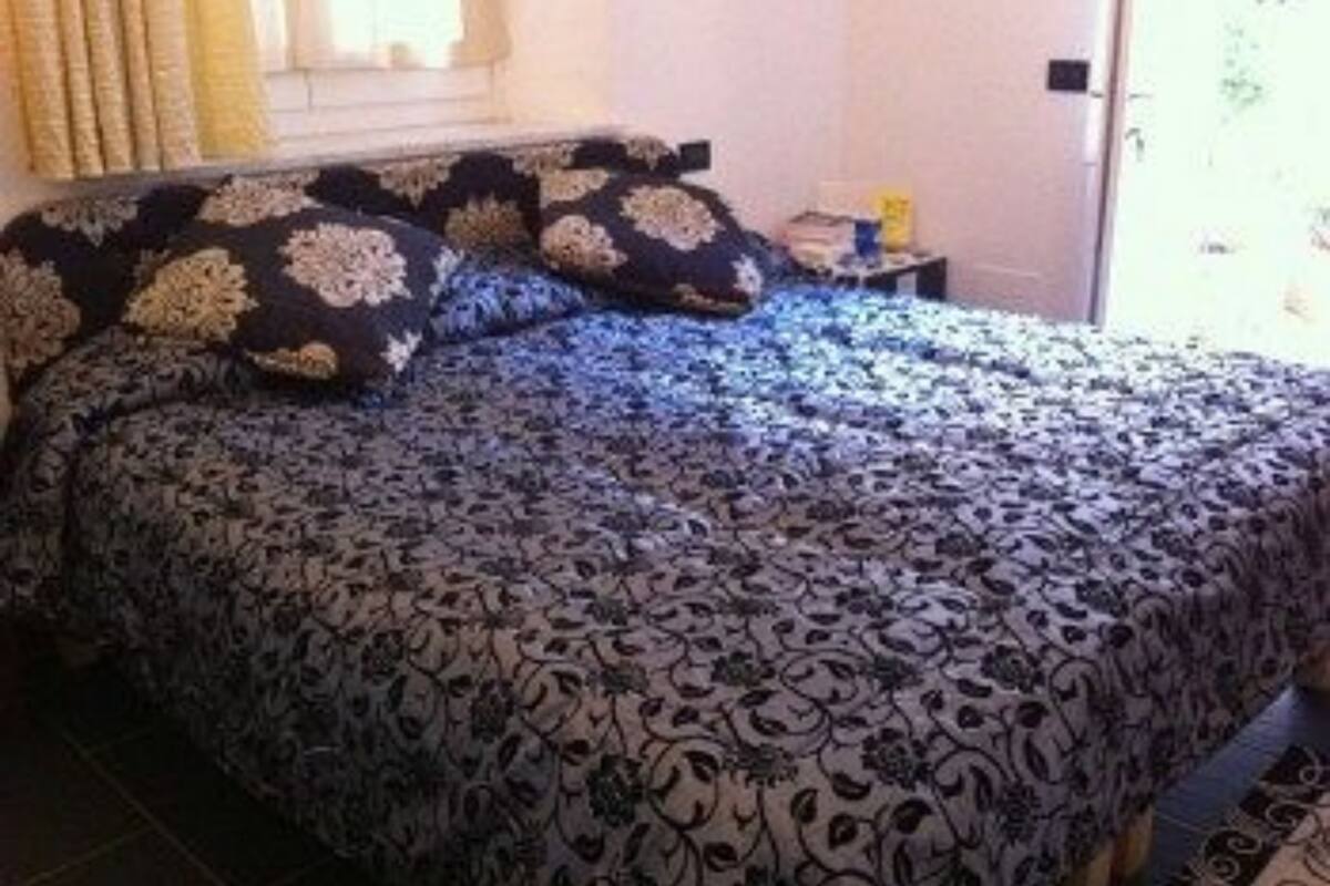 Una cama con una colcha estampada y almohadas a juego, una mesita de noche con lámpara, y una ventana con cortinas.