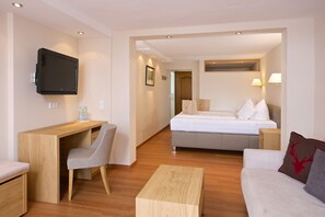 Une chambre d’hôtel moderne avec un bureau en bois, une chaise grise, un lit avec une literie blanche, un canapé blanc et une petite table en bois.