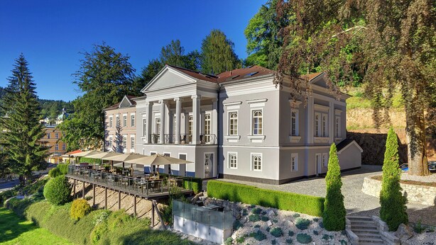 En stor byggnad i två våningar med en klassisk arkitektur, med ett takförsett utomhusområde med bord och parasoller, omgiven av välskötta trädgårdar och en stensatt gångväg som leder till entrén.